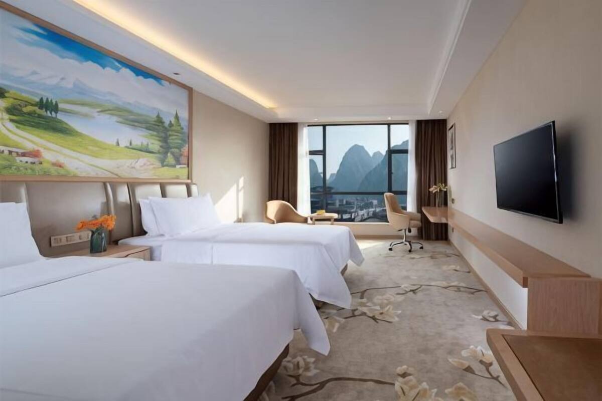 A hotel room with two beds, a large painting, a TV, and a view of mountains.