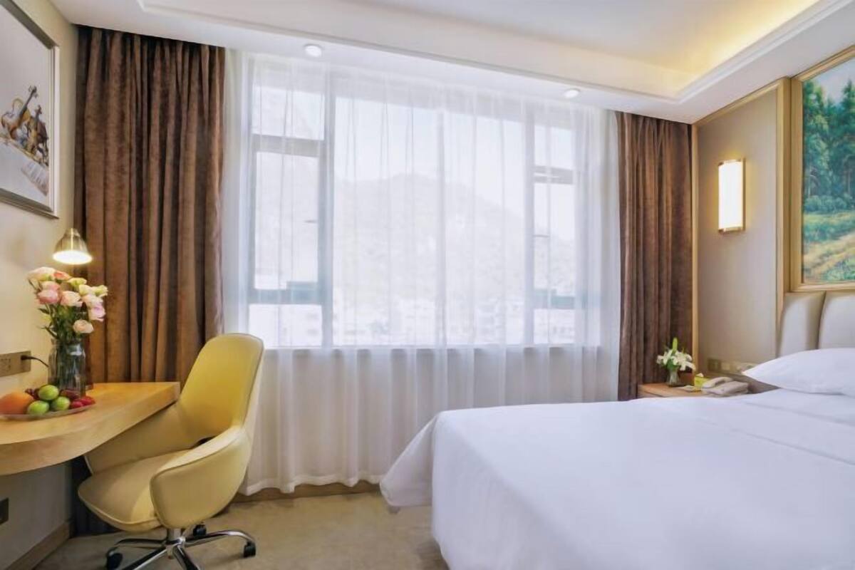 A hotel room with a bed, a desk with a chair, a vase of flowers, and a painting on the wall.