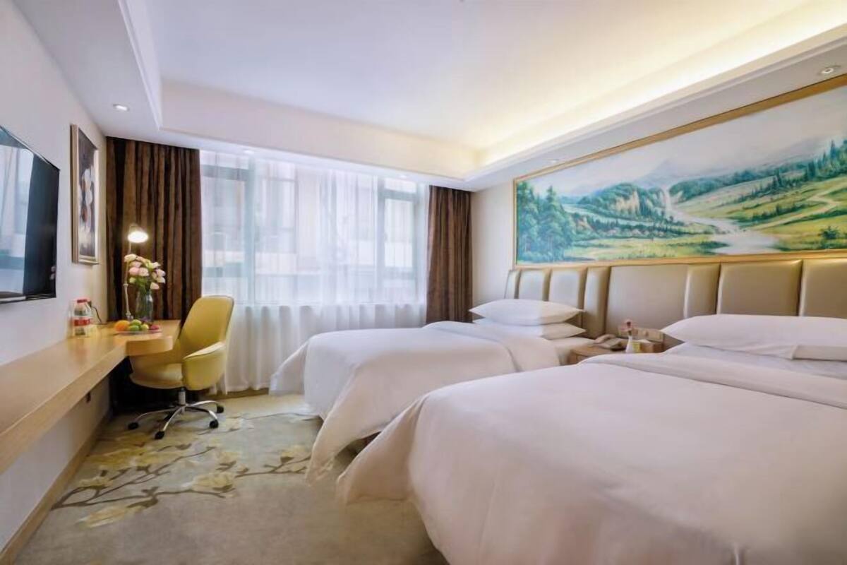 A hotel room with two beds, a desk with a chair, a painting on the wall, and a window with curtains.