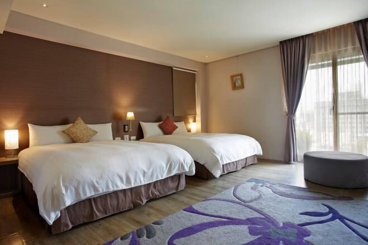 A hotel room with two beds, a round ottoman, a large window with curtains, and a patterned rug.