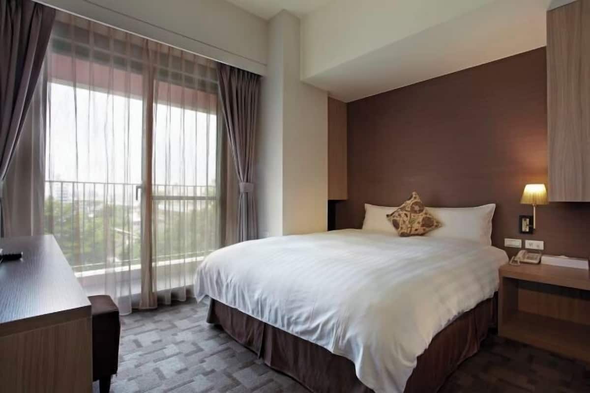 A hotel room with a bed, a desk, a chair, a bedside table, a lamp, and a view through a window with curtains.