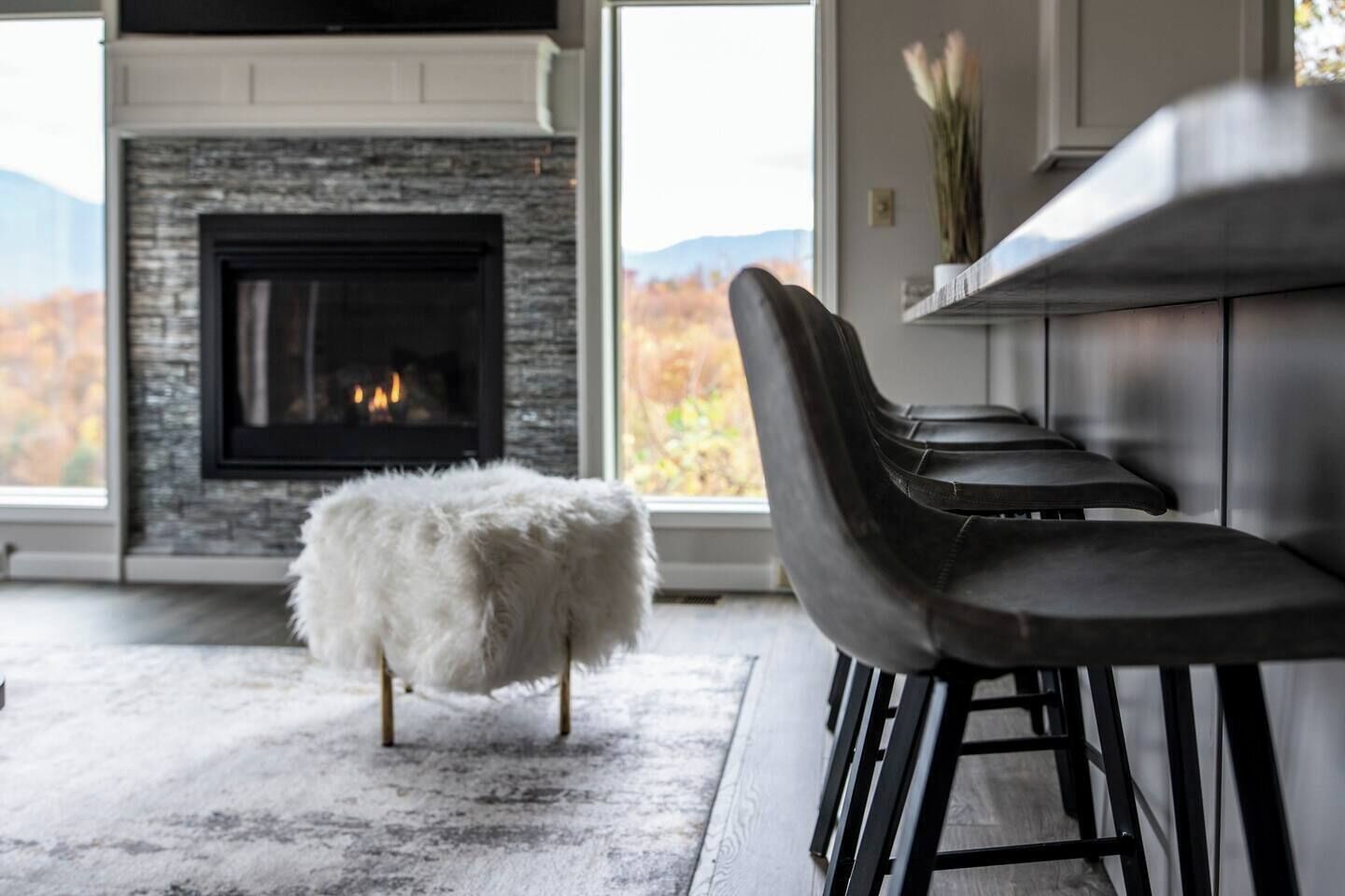 A modern living room with a fireplace, a white fluffy ottoman, and black bar stools.
