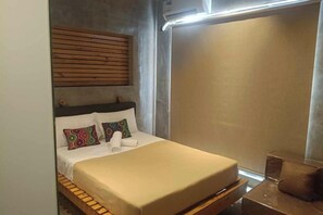 A bed with a wooden headboard, a bedside table, and a window with blinds. - Ramos Hello Stay Cebu (Cebu City)