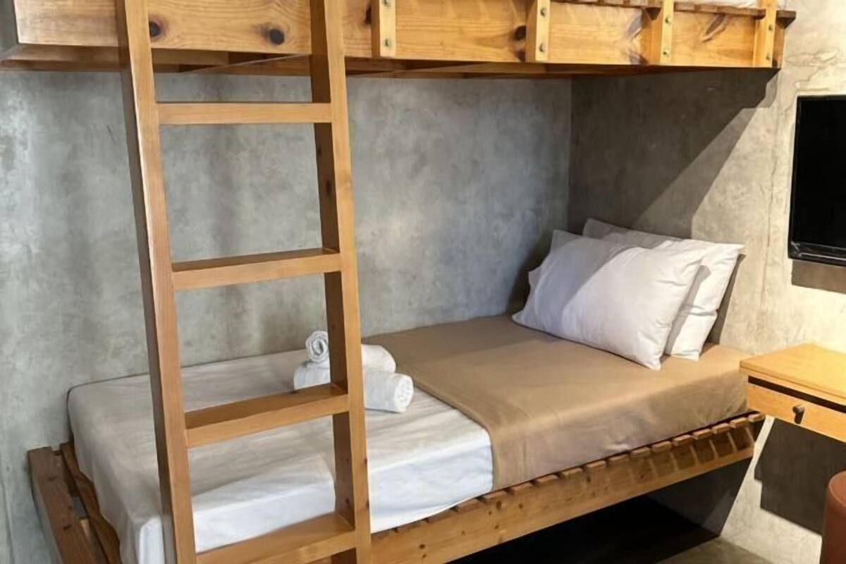 A bunk bed with a wooden ladder, a single bed with a white mattress and pillow, and a small wooden nightstand with a lamp.