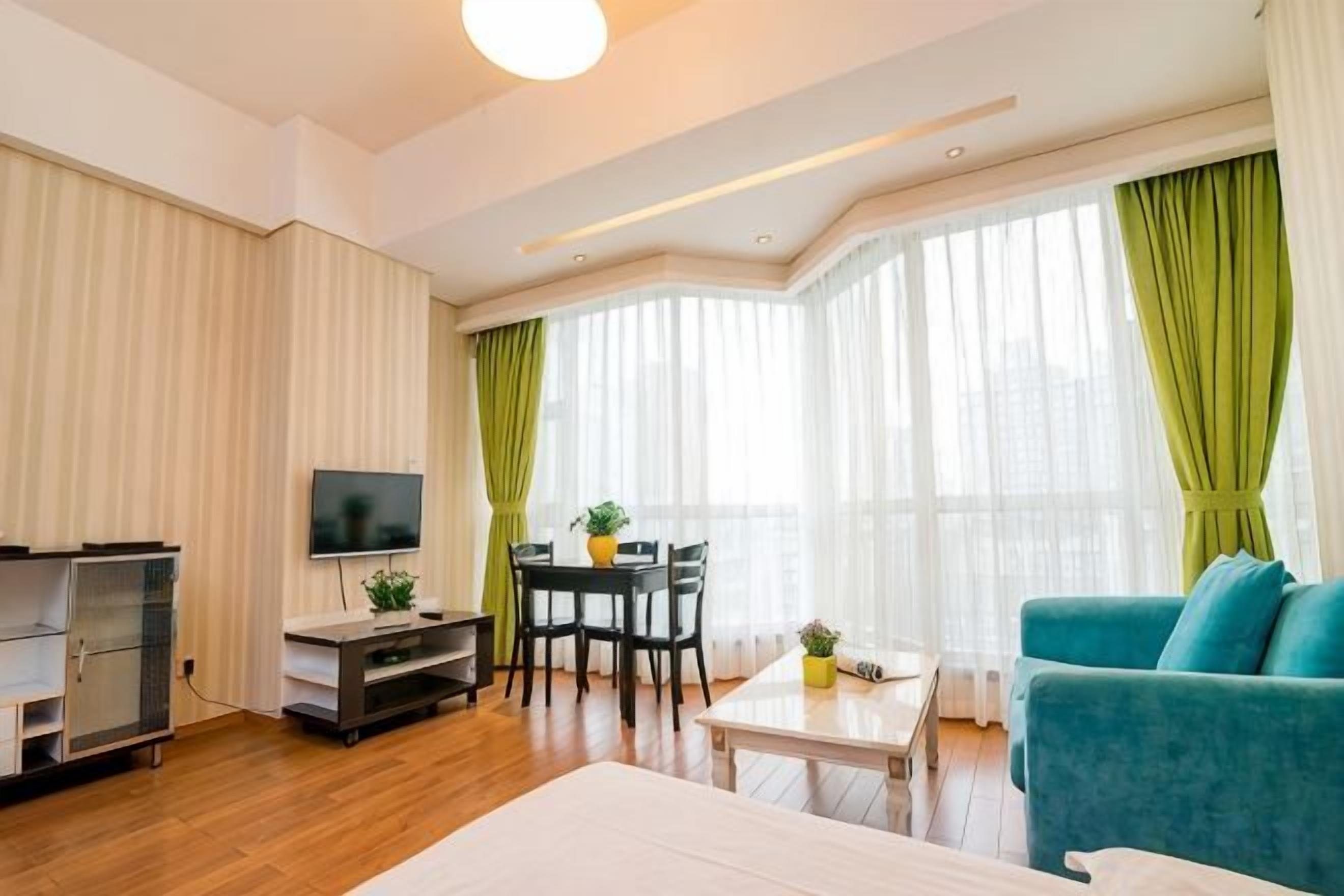 A hotel room with a bed, a TV, a dining table with chairs, a sofa, and large windows with curtains.