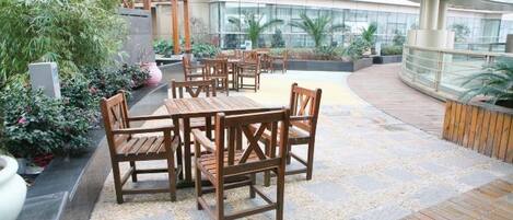 An outdoor seating area with wooden tables and chairs, surrounded by potted plants and a modern building in the background.