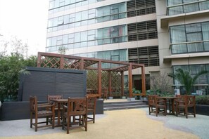Un espacio al aire libre con mobiliario de madera, una estructura cubierta y un edificio con grandes ventanales al fondo.
