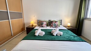 A hotel room with a bed, two nightstands, a green blanket, two pillows, and a window with curtains. - MONTE GORDO BEACH OCEAN VIEW 5 by HOMING (Monte Gordo)