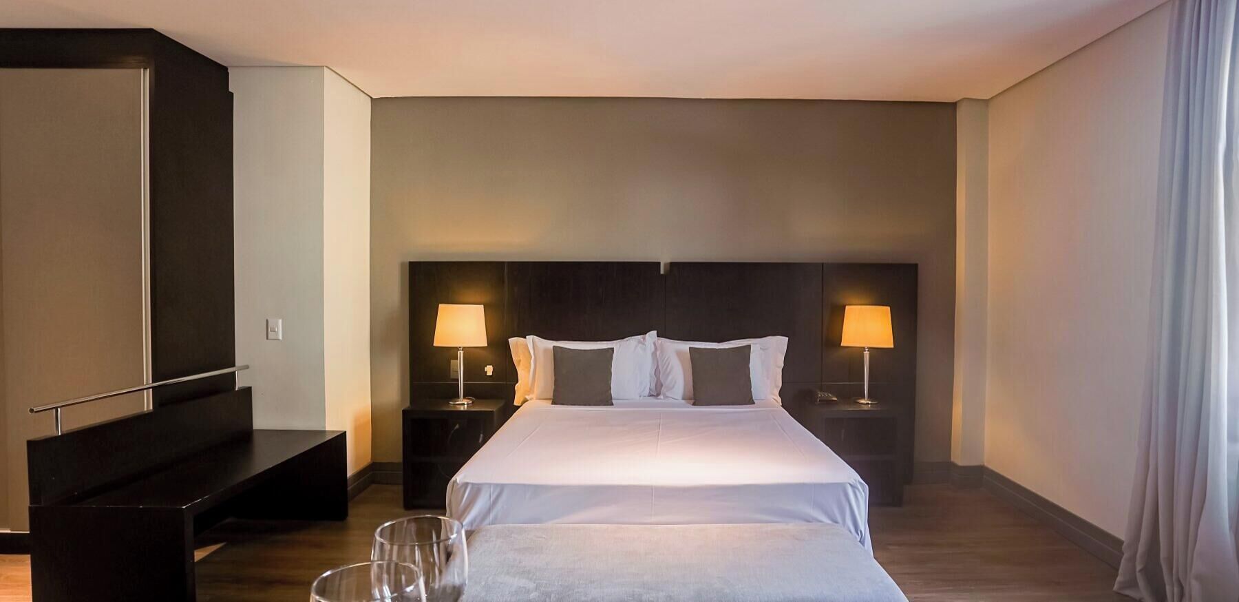 Ett hotellrum med en stor säng, två sängbord med lampor, en mörk sänggavel och en bänk vid fotändan av sängen.