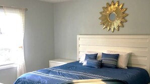Ein Schlafzimmer mit einem weißen Bett, blauer Bettwäsche, einem dekorativen Spiegel im Sonnenstrahl-Design und einem Fenster mit Vorhängen.
