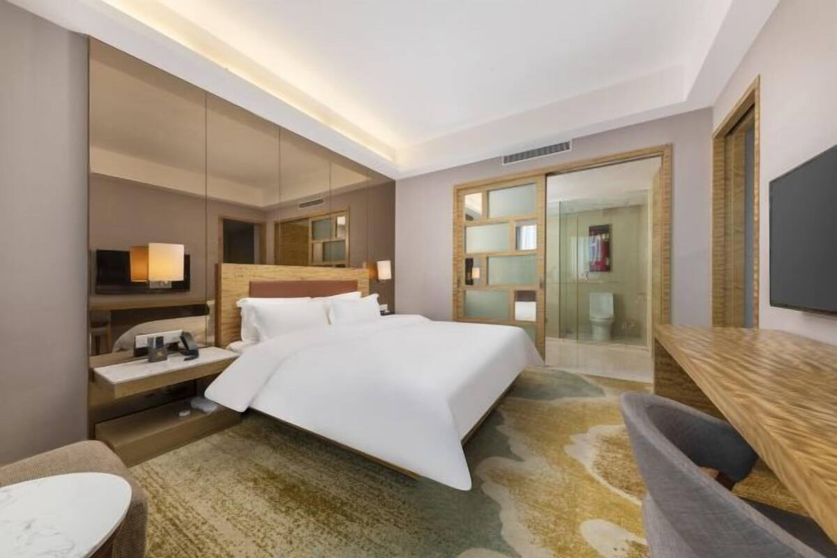 A hotel room with a bed, bedside table, lamp, TV, and a large mirror.