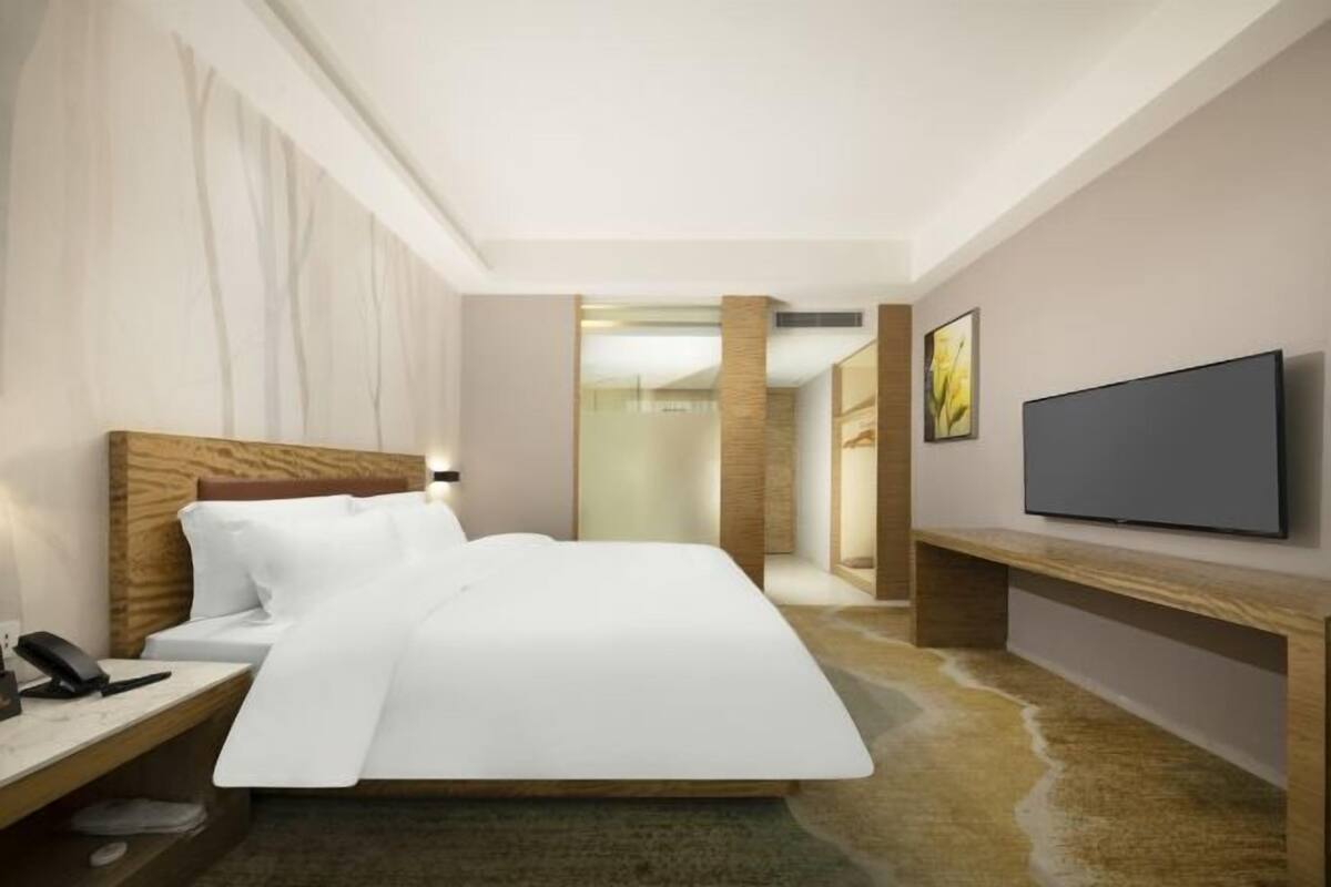 A modern hotel room with a large bed, a bedside table with a phone, a flat-screen TV mounted on the wall, and a wooden bench.