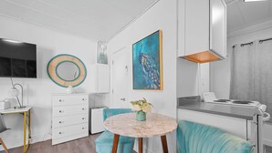 A modern kitchen with a white countertop, a stove, and a sink. There is a round table with a vase of flowers and two blue chairs. A painting of a peacock is on the wall, and a television is mounted above the counter. - Chic City Retreat 93A Sleeps 2 (Columbus)