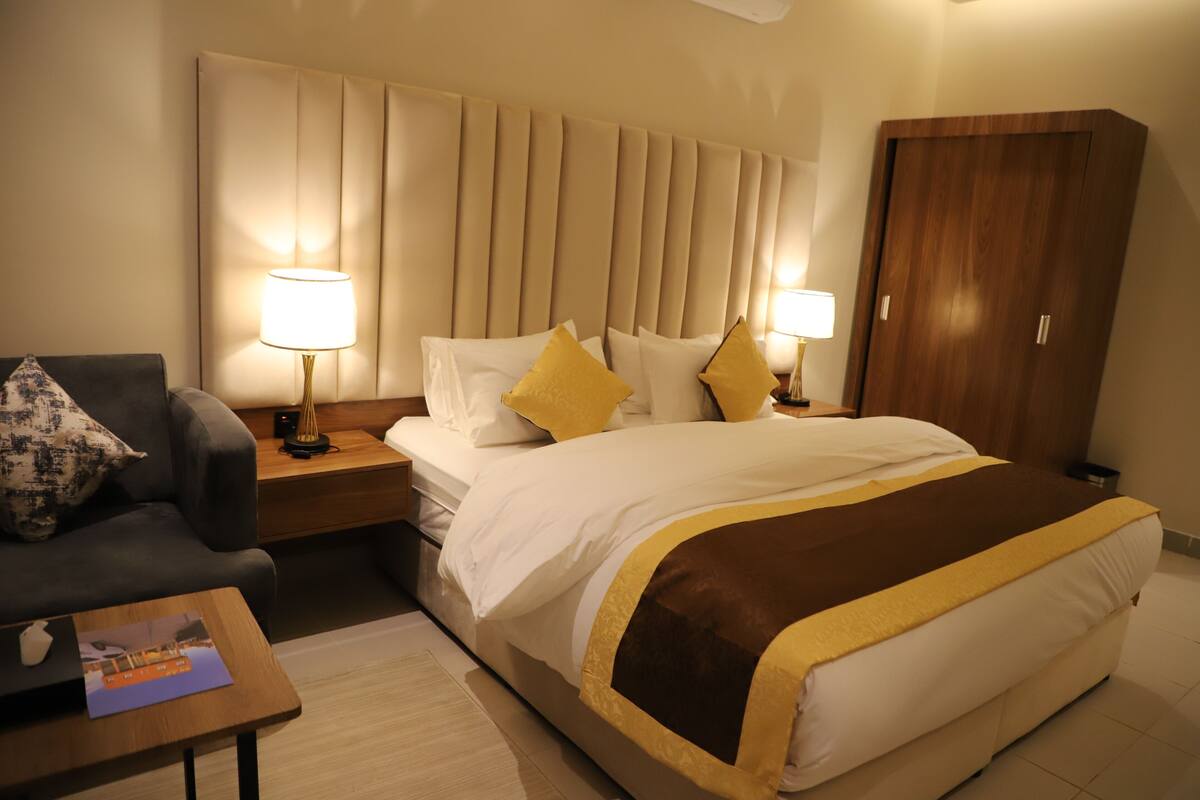 A hotel room with a large bed, two bedside lamps, a wooden nightstand, a dark armchair, and a small table with a magazine.