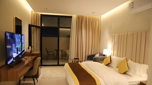 A hotel room with a bed, a desk with a TV, a chair, a lamp, and a sliding glass door leading to a balcony. - Afal Serviced Apartments - Al-Munsiyah (Riyadh)