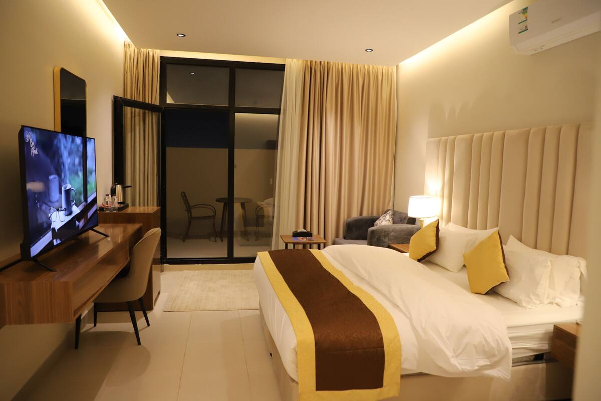 A hotel room with a bed, a desk with a TV, a chair, a lamp, and a sliding glass door leading to a balcony.