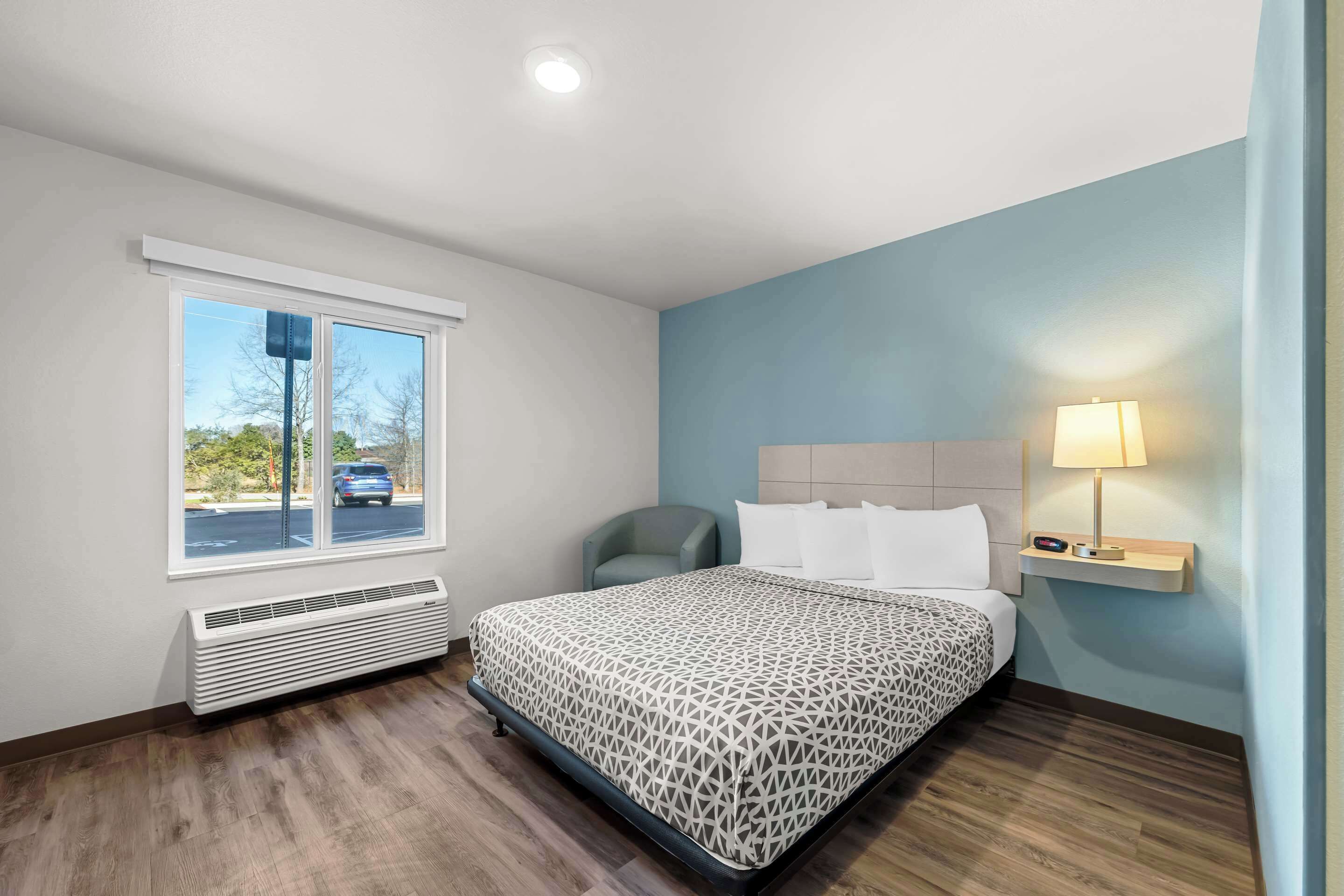 Une chambre d’hôtel avec un lit, une chaise, une fenêtre, un radiateur et une table de chevet avec une lampe.