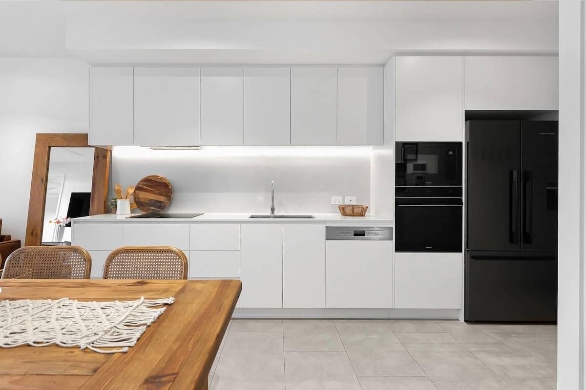 Una cocina moderna con armarios blancos, un refrigerador negro y un comedor con mesa de madera y sillas de mimbre.