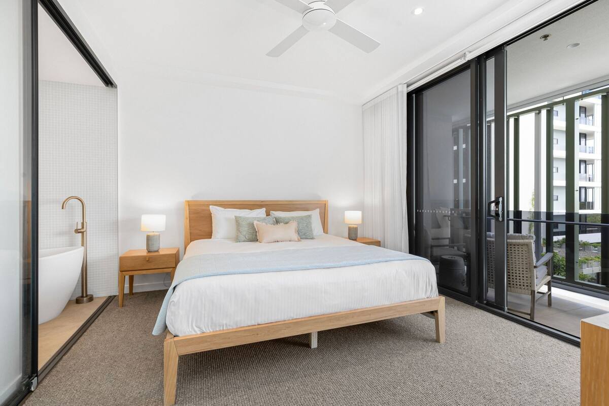 Relax in a spacious room with a large bed, wooden nightstand, and sliding glass door leading to a balcony with a city view.