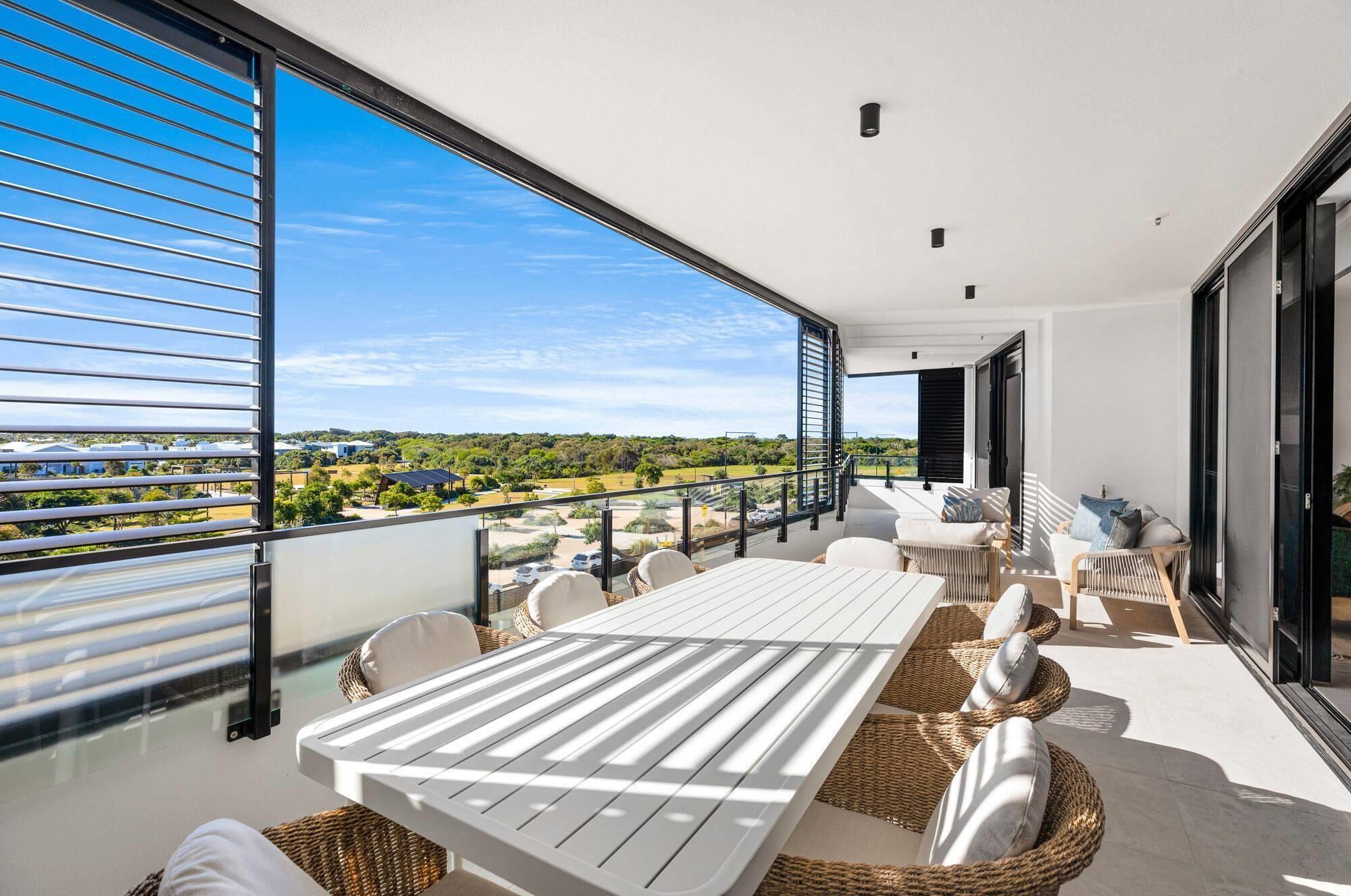 A balcony with a white table and wicker chairs, overlooking a residential area with houses and greenery.