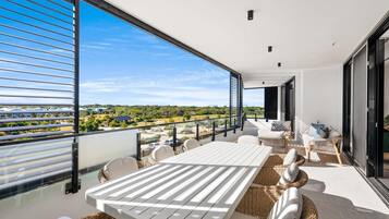 A balcony with a white table and wicker chairs, overlooking a residential area with houses and greenery.