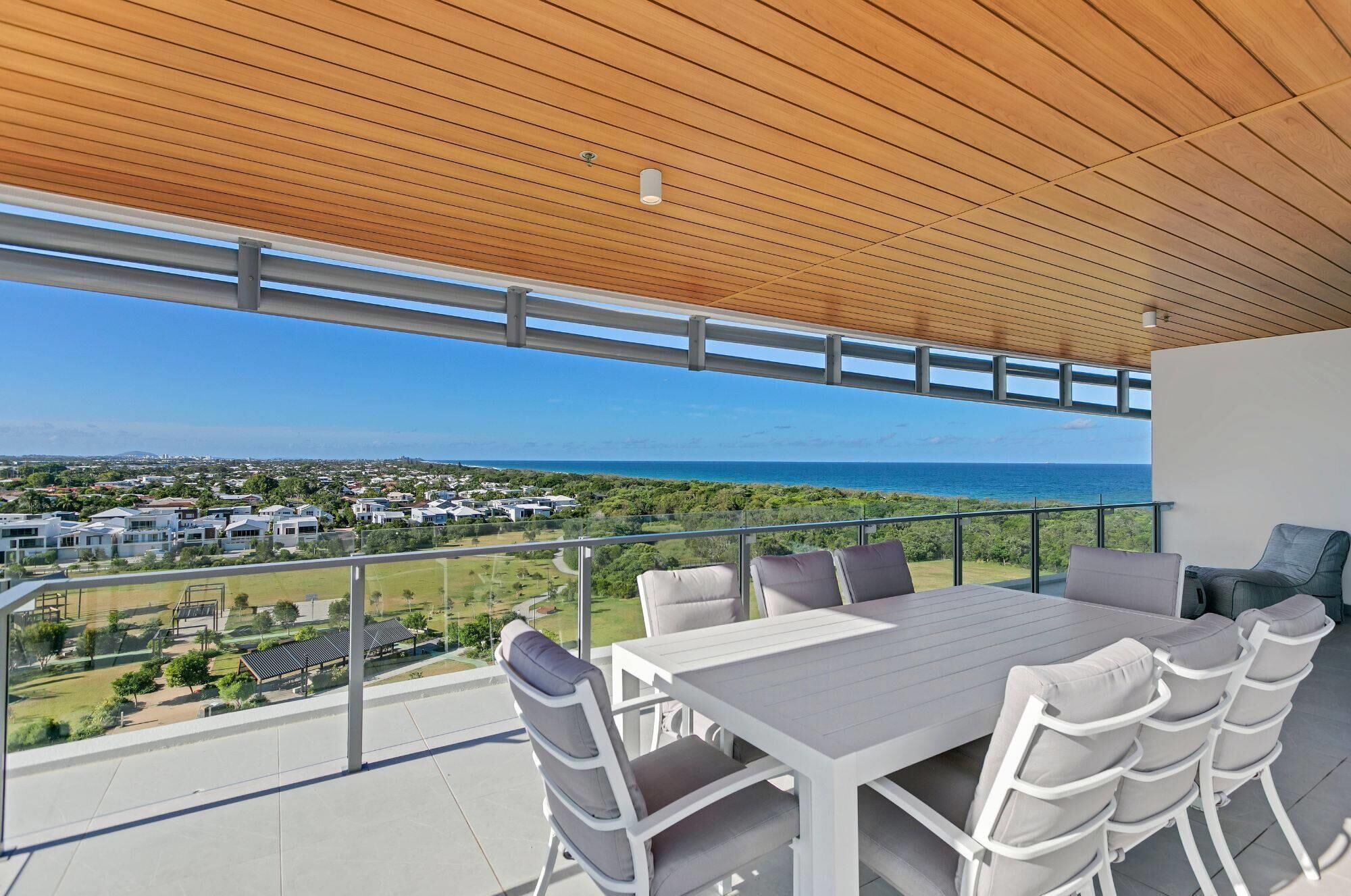 A balcony with a wooden ceiling, white outdoor furniture, and a view of the ocean and residential area.