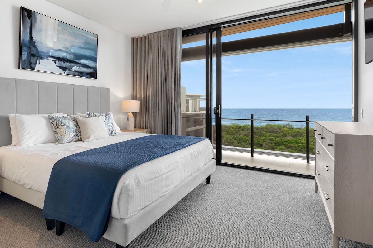 Una habitación de hotel moderna con una cama grande, una mesita de noche, un aparador y vista al océano.