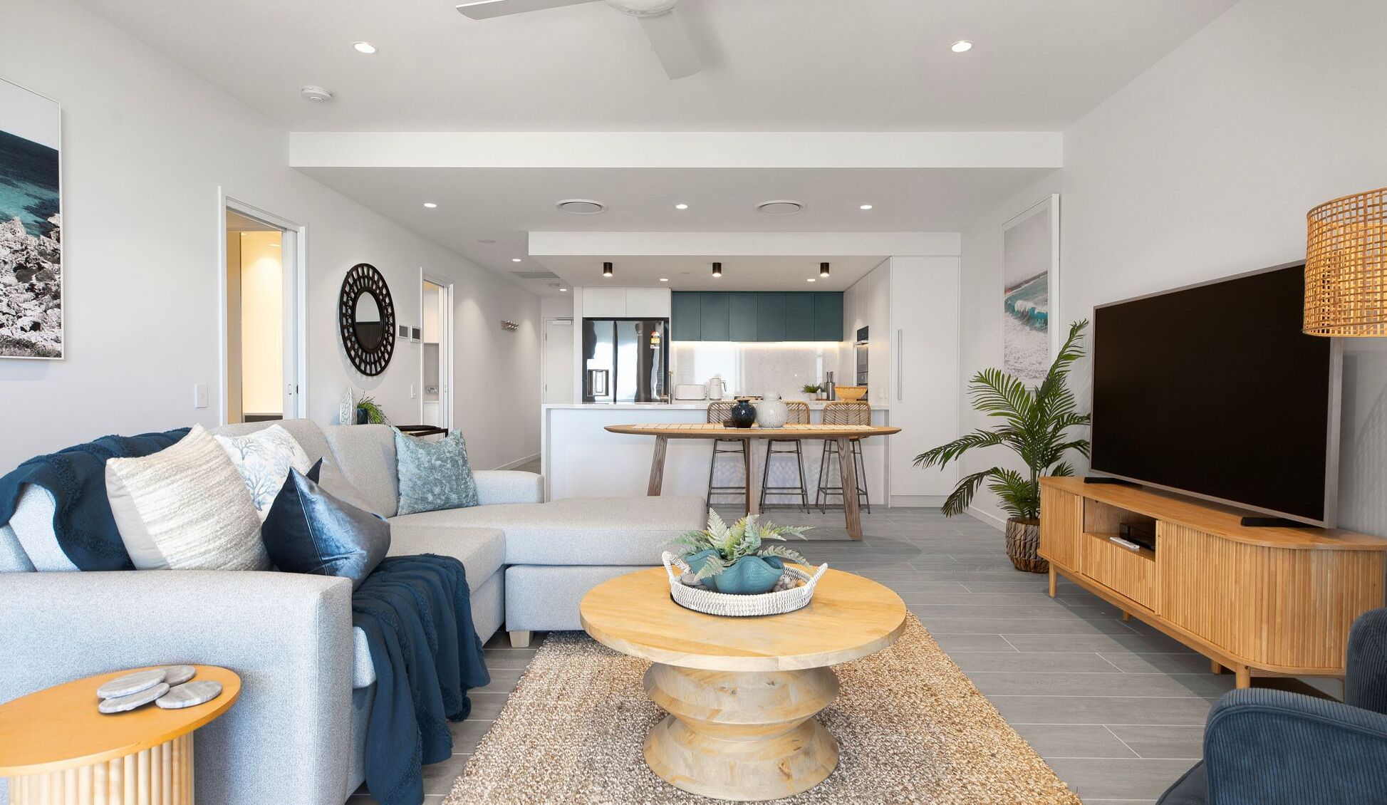 A modern living room with a sofa, coffee table, and TV. There is a kitchen in the background with a dining area.