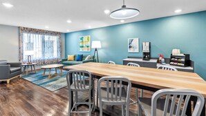 A modern living room with a wooden dining table, grey chairs, a sofa, and a coffee machine. - WoodSpring Suites St Peters-OFallon (St. Peters)