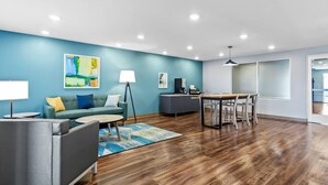 A modern living room with a blue accent wall, a gray sofa, a dining table with chairs, and a coffee table. - WoodSpring Suites St Peters-OFallon (St. Peters)