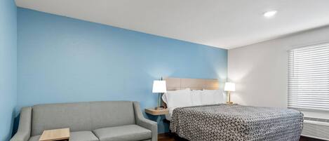 Una habitación de hotel con un sofá gris, una cama con colcha estampada, una mesita de noche con lámpara y una ventana con persianas.
