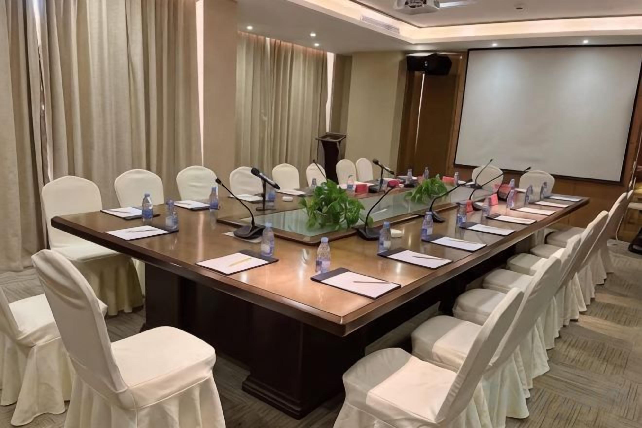 A conference room with a long table, chairs, microphones, and a projector screen.