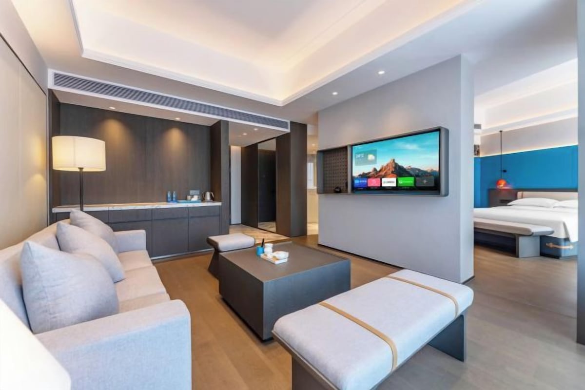 A modern hotel room with a sofa, a coffee table, a TV mounted on the wall, and a bed.