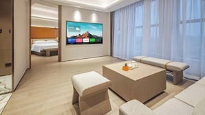 A modern hotel room with a large flat-screen TV displaying a mountain landscape, a comfortable sofa, a coffee table, and a neatly made bed. - Rufu Hotel (Ganzhou)