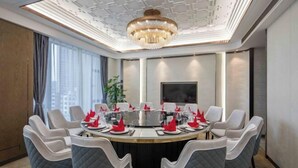 Un salón de conferencias con una mesa redonda, sillas blancas y grises, una lámpara de araña y una pantalla grande.