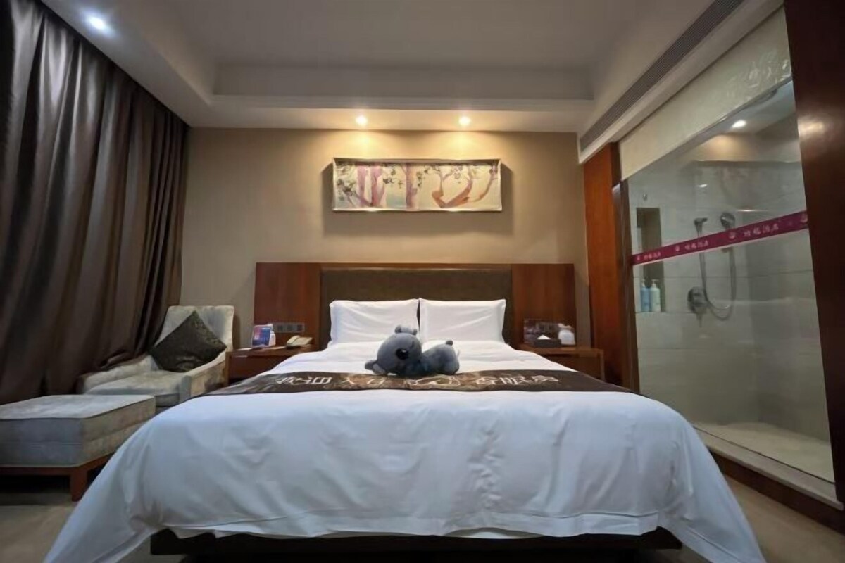 A hotel room with a large bed, a chair, a nightstand, a painting on the wall, and a glass shower enclosure.