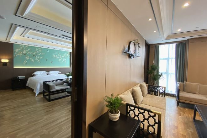 Una habitación de hotel moderna con piso de madera, una cama con ropa blanca, un sofá, una mesita con una planta y un reloj de pared.