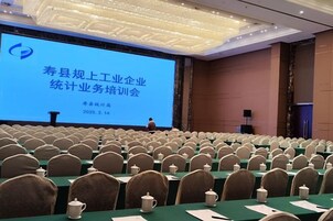 Un salón de conferencias con filas de sillas y mesas, una pantalla grande mostrando texto, y un atril.