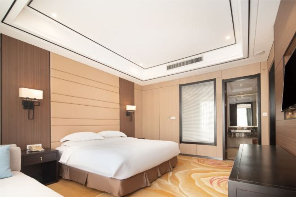 Une chambre d’hôtel comprenant un lit, des tables de chevet, un bureau, un téléviseur à écran plat et une grande fenêtre avec des stores.