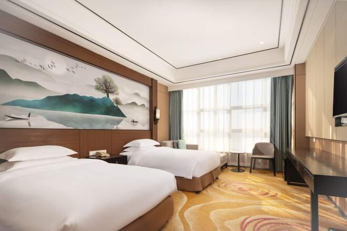 Ein Hotelzimmer mit zwei Betten, einem Schreibtisch, einem Sessel, einem Fernseher und einem großen Gemälde an der Wand.
