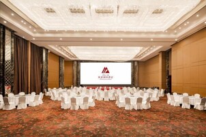 Un salón de conferencias con filas de mesas y sillas, una pantalla grande al frente y piso alfombrado.