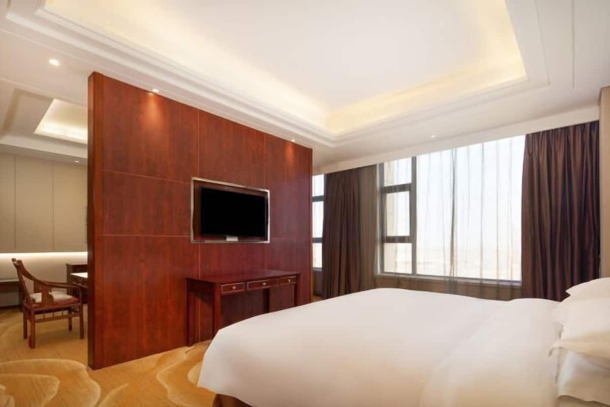 Ein Hotelzimmer mit einem großen Bett, einem an einer Holzplatte montierten Fernseher, einem Schreibtisch mit Stuhl und einem großen Fenster mit Vorhängen.