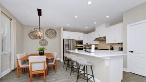 A modern kitchen with white cabinets, a central island, bar stools, and a dining table with chairs. - Inviting Home near Disney with Waterpark & Resort Access at Encore Resort -. (Kissimmee)