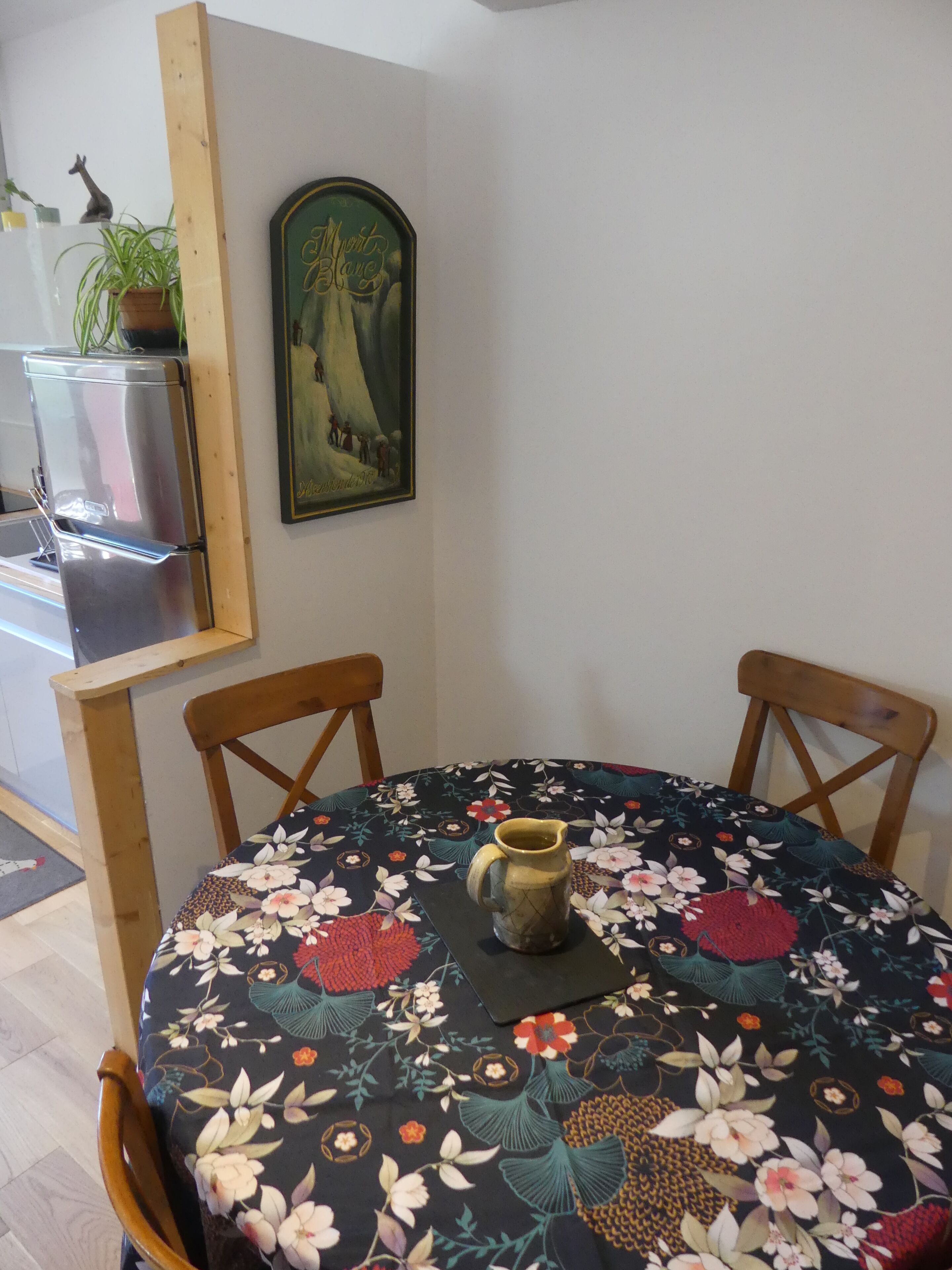 Un coin repas avec une table ronde recouverte d’une nappe à motifs floraux, deux chaises en bois, un pichet en céramique, un tableau encadré accroché au mur et une plante en pot posée sur le comptoir.