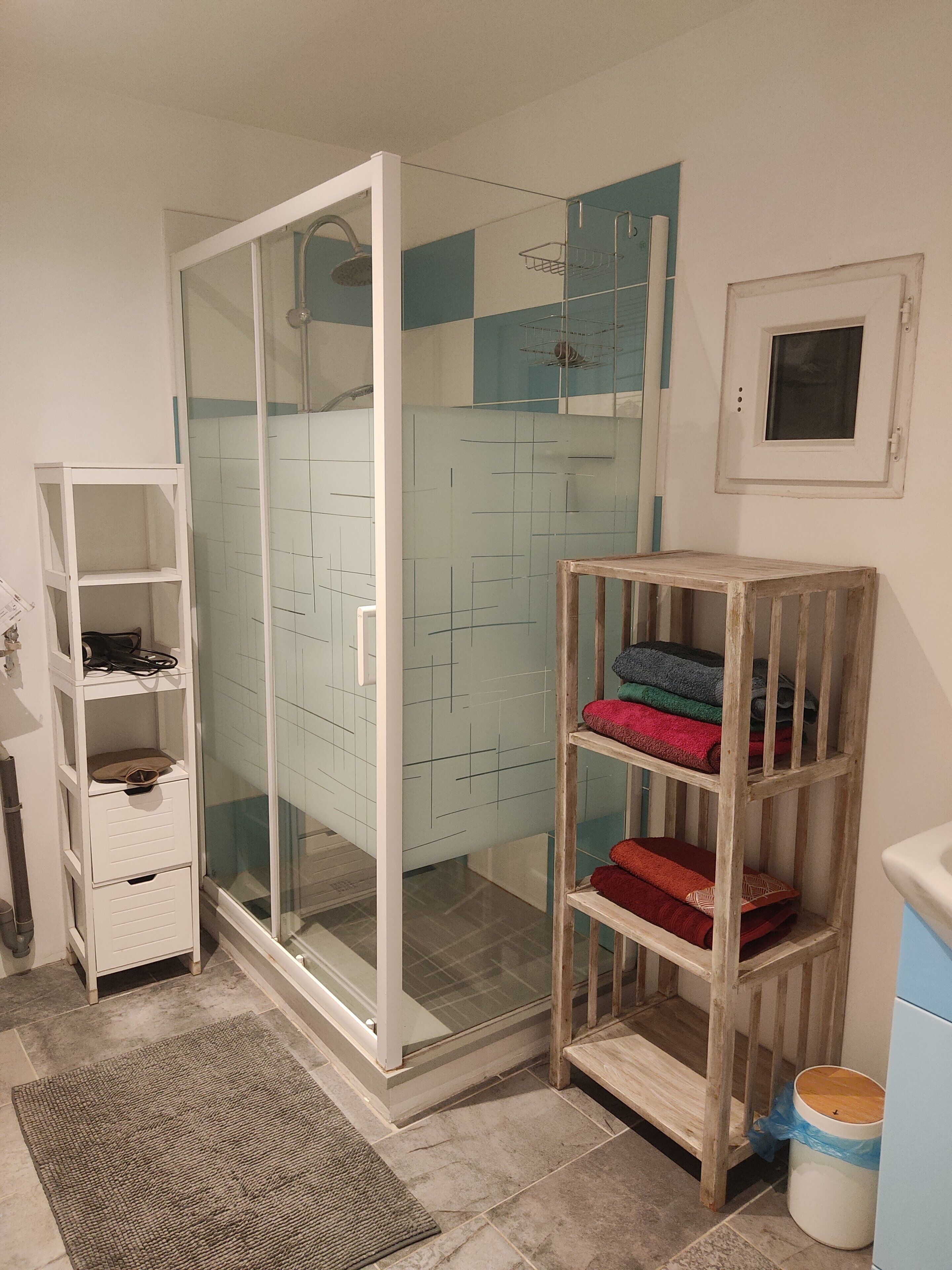 A bathroom with a glass shower enclosure, a wooden shelf with towels, a white cabinet, and a tiled floor.