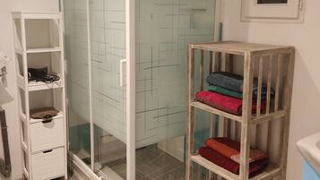 A bathroom with a glass shower enclosure, a wooden shelf with towels, a white cabinet, and a tiled floor.
