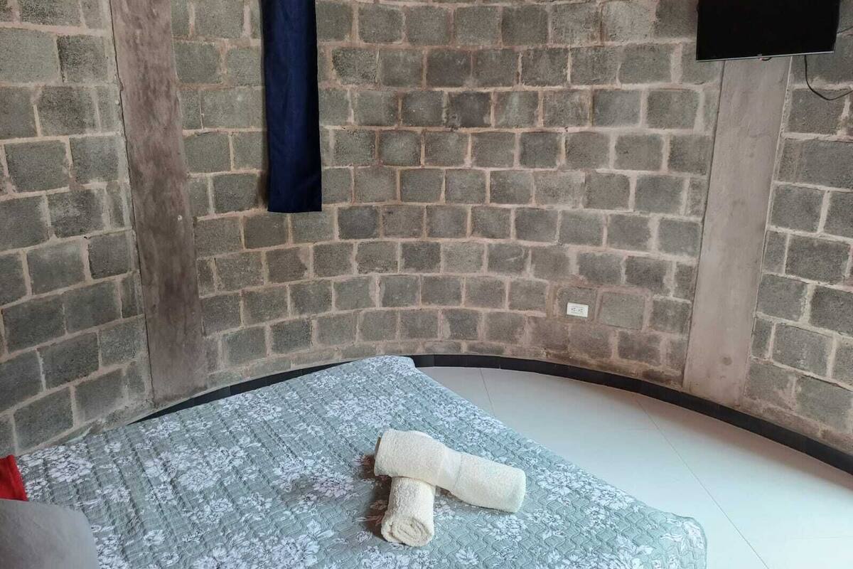 A room with a brick wall, a flat-screen TV mounted on the wall, and a bed with a floral-patterned mattress and a rolled-up white towel.