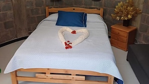 Ein Bett mit Holzrahmen, eine weiße gesteppte Matratze, ein blaues Kopfkissen und ein herzförmiges Handtuch mit roten Blumen. Dazu ein Nachttisch aus Holz mit einer Vase getrockneter Blumen.