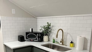 Et køkken med hvidt flisebaggrund, en stålvask, en sort mikroovn, en hvid vase med en plante, en gul sæbedispenser og en rulle køkkenrulle.