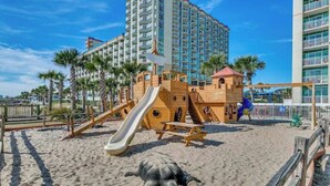 Un’area giochi sulla spiaggia con scivolo e strutture per arrampicarsi, circondata da palme e un alto edificio di un hotel.